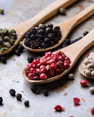 Auf einer Metalloberfläche liegen vier Holzlöffel. Jeder Löffel ist mit einer anderen Pfeffersorte gefüllt: grüne, schwarze, rote und weiße Pfefferkörner.