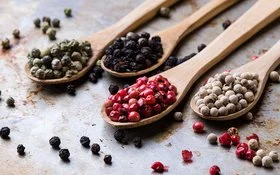 Auf einer Metalloberfläche liegen vier Holzlöffel. Jeder Löffel ist mit einer anderen Pfeffersorte gefüllt: grüne, schwarze, rote und weiße Pfefferkörner.