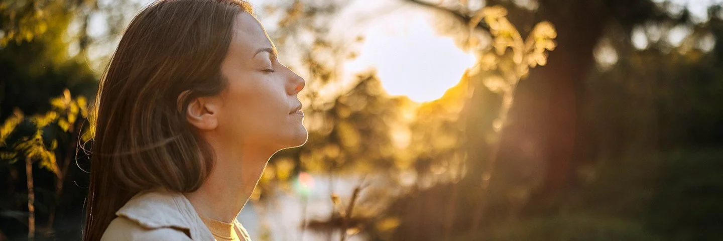 Eine Frau sitzt mit geschlossenen Augen in der Natur. Die tief stehende Sonne strahlt ihr ins Gesicht. Sie wirkt entspannt.