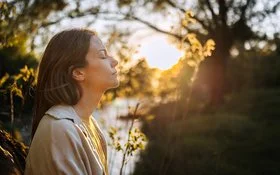 Eine Frau sitzt mit geschlossenen Augen in der Natur. Die tief stehende Sonne strahlt ihr ins Gesicht. Sie wirkt entspannt.
