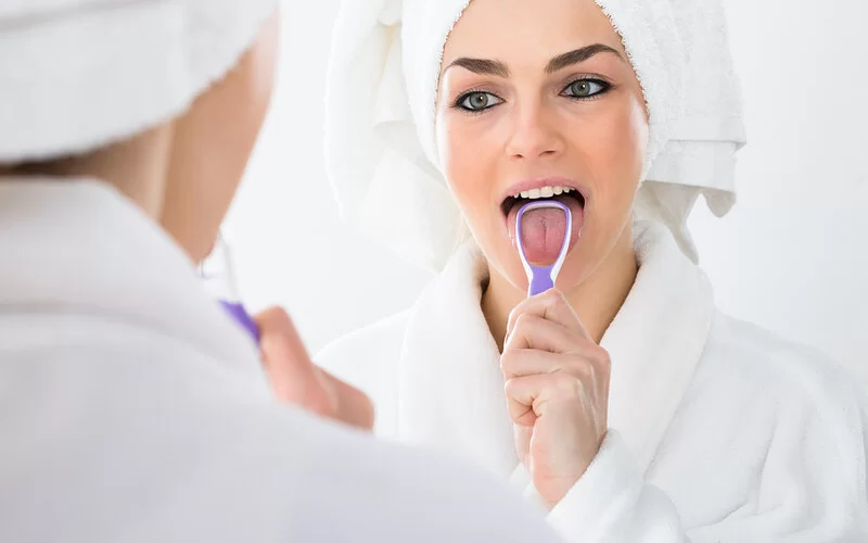 Frau steht vor dem Badezimmerspiegel und reinigt ihre Zunge mit einem Zungenreiniger.