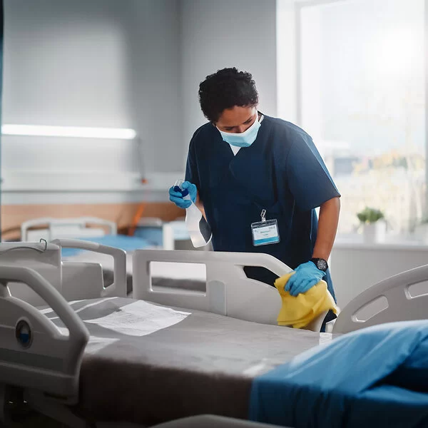 Eine weibliche Pflegekraft in einem Krankenzimmer reinigt ein Pflegebett mit Desinfektionsmittel.