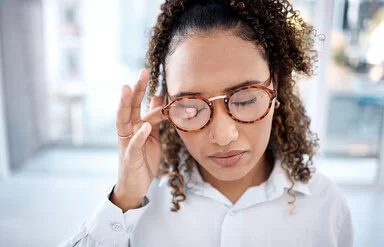 Eine junge Frau mit hochgesteckten langen, braunen Locken reibt sich mit den Fingern ein Auge. Sie trägt eine Brille und hält beide Augen geschlossen.