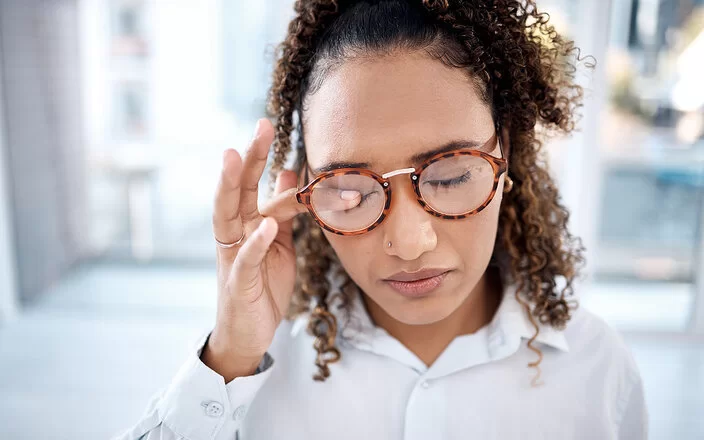 Eine junge Frau mit hochgesteckten langen, braunen Locken reibt sich mit den Fingern ein Auge. Sie trägt eine Brille und hält beide Augen geschlossen.