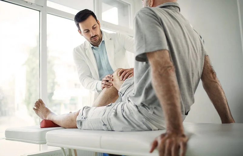 Ein Arzt in weißem Kittel untersucht das Knie eines älteren Mannes, der auf einer Untersuchungsliege sitzt.