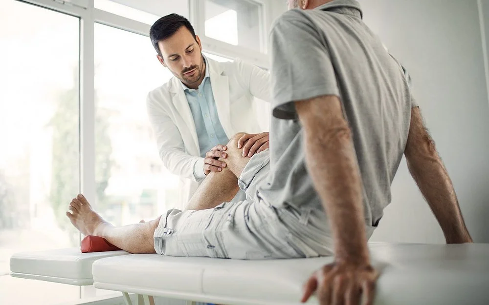 Ein Arzt in weißem Kittel untersucht das Knie eines älteren Mannes, der auf einer Untersuchungsliege sitzt.