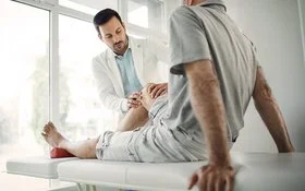 Ein Arzt in weißem Kittel untersucht das Knie eines älteren Mannes, der auf einer Untersuchungsliege sitzt.