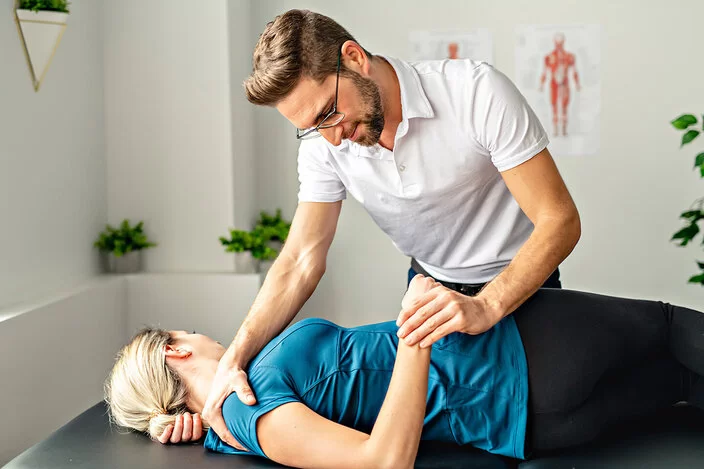 Eine Frau liegt mit dem Rücken auf einer Liege. Ein Osteopath beugt sich über sie und hält dabei mit seiner rechten Hand ihre rechte Schulter, mit seiner linken Hand hält er ihre rechte Hand und bewegt ihren Unterarm.