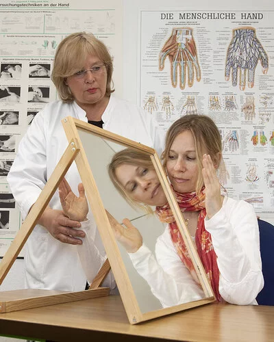In einem Behandlungszimmer führt eine Physiotherapeutin mit einer jungen Patientin eine Spiegeltherapie durch. Die Patientin sitzt an einem Tisch, auf dem rechtwinklig zur Längsseite ein Spiegel aufgestellt ist. Die Therapeutin hält die rechte Hand der Patientin hinter dem Spiegel fest. Die linke Hand bewegt die junge Frau vor dem Spiegel und betrachtet dabei das Spiegelbild.