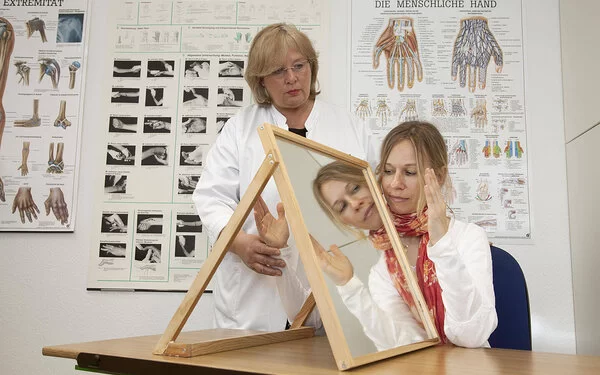 In einem Behandlungszimmer führt eine Physiotherapeutin mit einer jungen Patientin eine Spiegeltherapie durch. Die Patientin sitzt an einem Tisch, auf dem rechtwinklig zur Längsseite ein Spiegel aufgestellt ist. Die Therapeutin hält die rechte Hand der Patientin hinter dem Spiegel fest. Die linke Hand bewegt die junge Frau vor dem Spiegel und betrachtet dabei das Spiegelbild.
