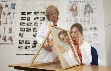 In einem Behandlungszimmer führt eine Physiotherapeutin mit einer jungen Patientin eine Spiegeltherapie durch. Die Patientin sitzt an einem Tisch, auf dem rechtwinklig zur Längsseite ein Spiegel aufgestellt ist. Die Therapeutin hält die rechte Hand der Patientin hinter dem Spiegel fest. Die linke Hand bewegt die junge Frau vor dem Spiegel und betrachtet dabei das Spiegelbild.