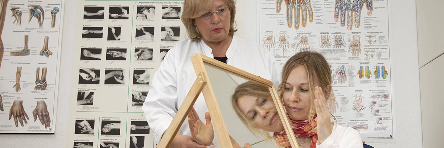 In einem Behandlungszimmer führt eine Physiotherapeutin mit einer jungen Patientin eine Spiegeltherapie durch. Die Patientin sitzt an einem Tisch, auf dem rechtwinklig zur Längsseite ein Spiegel aufgestellt ist. Die Therapeutin hält die rechte Hand der Patientin hinter dem Spiegel fest. Die linke Hand bewegt die junge Frau vor dem Spiegel und betrachtet dabei das Spiegelbild.