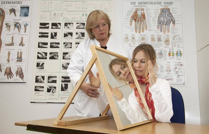 In einem Behandlungszimmer führt eine Physiotherapeutin mit einer jungen Patientin eine Spiegeltherapie durch. Die Patientin sitzt an einem Tisch, auf dem rechtwinklig zur Längsseite ein Spiegel aufgestellt ist. Die Therapeutin hält die rechte Hand der Patientin hinter dem Spiegel fest. Die linke Hand bewegt die junge Frau vor dem Spiegel und betrachtet dabei das Spiegelbild.