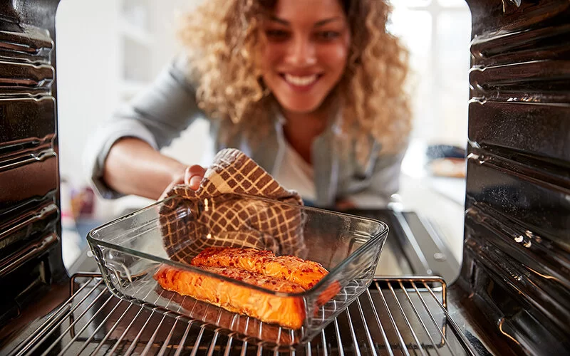 Eine Frau mit blonden, lockigen Haaren blickt lachend in einen Backofen. In der einen Hand hält sie mit einem Topflappen eine Auflaufform mit bereits gebräunten Lachsfilets.