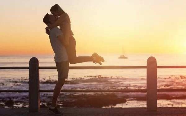  Ein Mann steht mit seiner Freundin bei Sonnenuntergang an einer Strandpromenade und hebt sie hoch, um sie zu küssen.