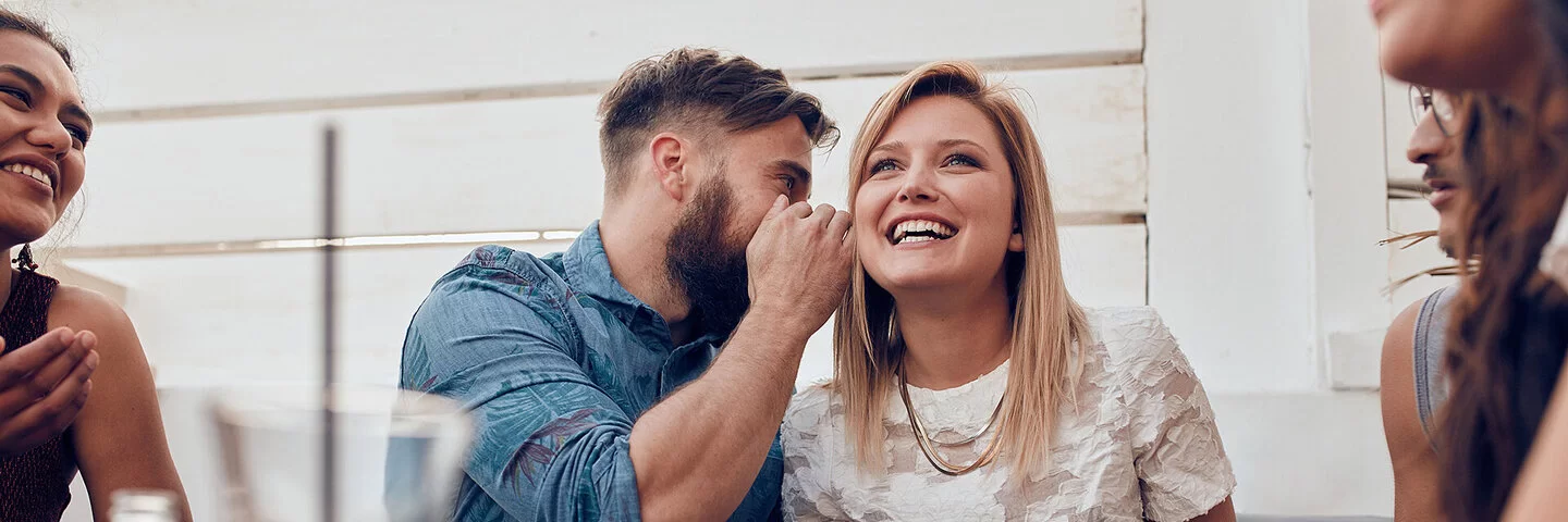 Eine Frau und ein Mann sitzen nebeneinander. Der Mann sagt der Frau etwas ins Ohr. 