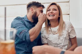 Eine Frau und ein Mann sitzen nebeneinander. Der Mann sagt der Frau etwas ins Ohr.