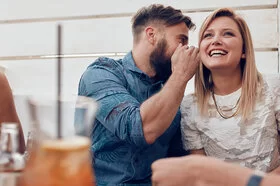 Eine Frau und ein Mann sitzen nebeneinander. Der Mann sagt der Frau etwas ins Ohr. 