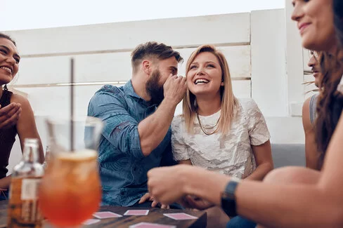 Eine Frau und ein Mann sitzen nebeneinander. Der Mann sagt der Frau etwas ins Ohr. 