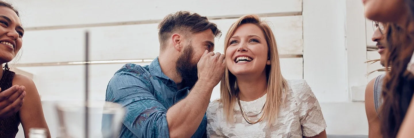 Eine Frau und ein Mann sitzen nebeneinander. Der Mann sagt der Frau etwas ins Ohr.