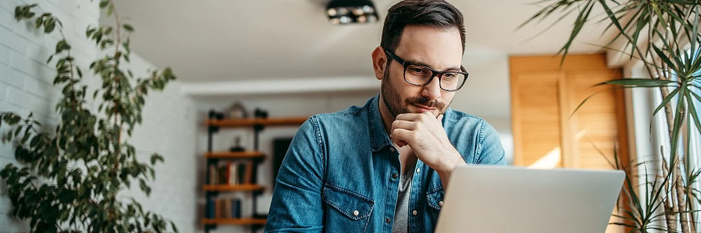 Ein Mann sitzt in einem Wohnzimmer an einem Tisch, vor ihm stehen eine Tasse und ein Laptop. Der Mann stützt sein Kinn auf seine linke Hand, er schaut interessiert auf das Display der Laptops.
