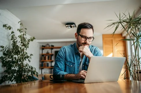 Ein Mann sitzt in einem Wohnzimmer an einem Tisch, vor ihm stehen eine Tasse und ein Laptop. Der Mann stützt sein Kinn auf seine linke Hand, er schaut interessiert auf das Display der Laptops.