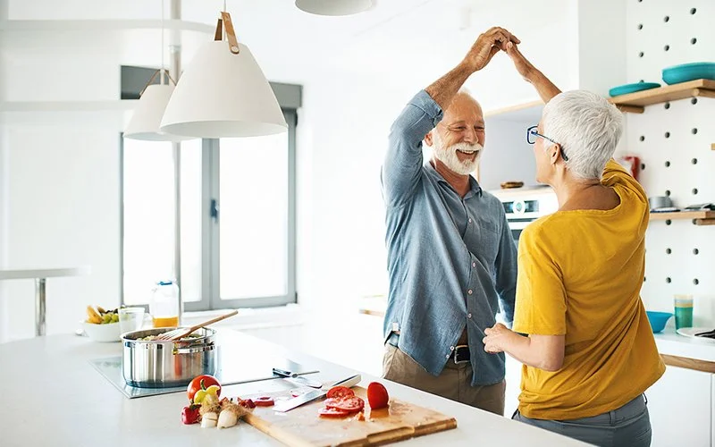 Ein glückliches älteres Paar tanzt beim gemeinsamen Kochen in einer hellen, modernen Küche.
