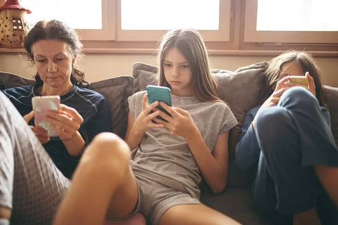 Eine Mutter sitzt lässig mit ihren beiden Töchtern, die im Teenageralter sind, auf dem Sofa im Wohnzimmer. Jede hat ein Smartphone in der Hand und blickt gebannt darauf. Sie nehmen keine Notiz voneinander.
