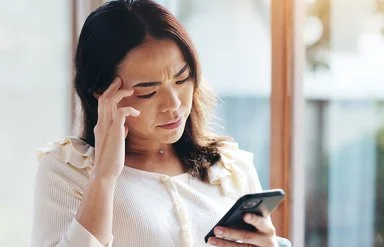 Frau steht am Fenster und blickt besorgt auf ihr Smartphone, während sie sich an die Stirn fasst.