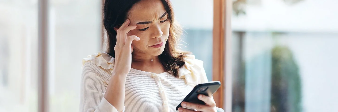 Frau steht am Fenster und blickt besorgt auf ihr Smartphone, während sie sich an die Stirn fasst.