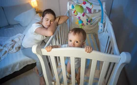 Junge müde Mutter schläft neben dem Kinderbett des Babys, während das Kind wach im Kinderbett steht.