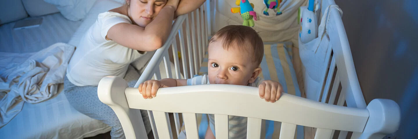 Junge müde Mutter schläft neben dem Kinderbett des Babys, während das Kind wach im Kinderbett steht.