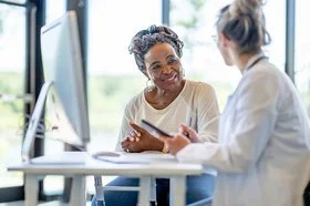 Eine Frau im mittleren Alter sitz an einem Schreibtisch mit einer Ärztin und die beiden unterhalten sich. 