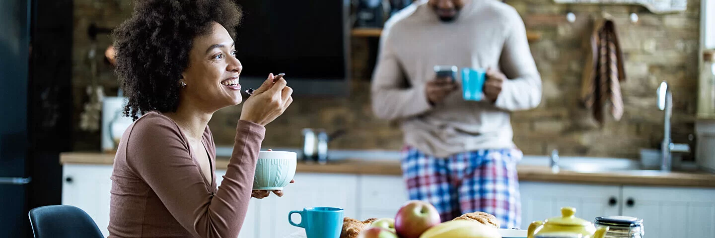 Eine Frau isst in der Küche Frühstück, ihr Freund steht im Hintergrund.