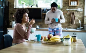 Eine Frau isst in der Küche Frühstück, ihr Freund steht im Hintergrund.