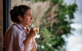 Eine Frau mit Kehlkopfentzündung steht vor einem Fenster, trinkt Tee und fasst sich an den Hals.