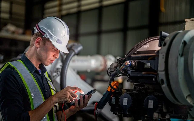 In einer Fabrikhalle steht ein Mann mit einem Schutzhelm vor einer Maschine. Er hält ein elektronisches Wartungs- oder Steuergerät in der linken Hand, blickt auf dessen Display und tippt mit der rechten Hand etwas ein.