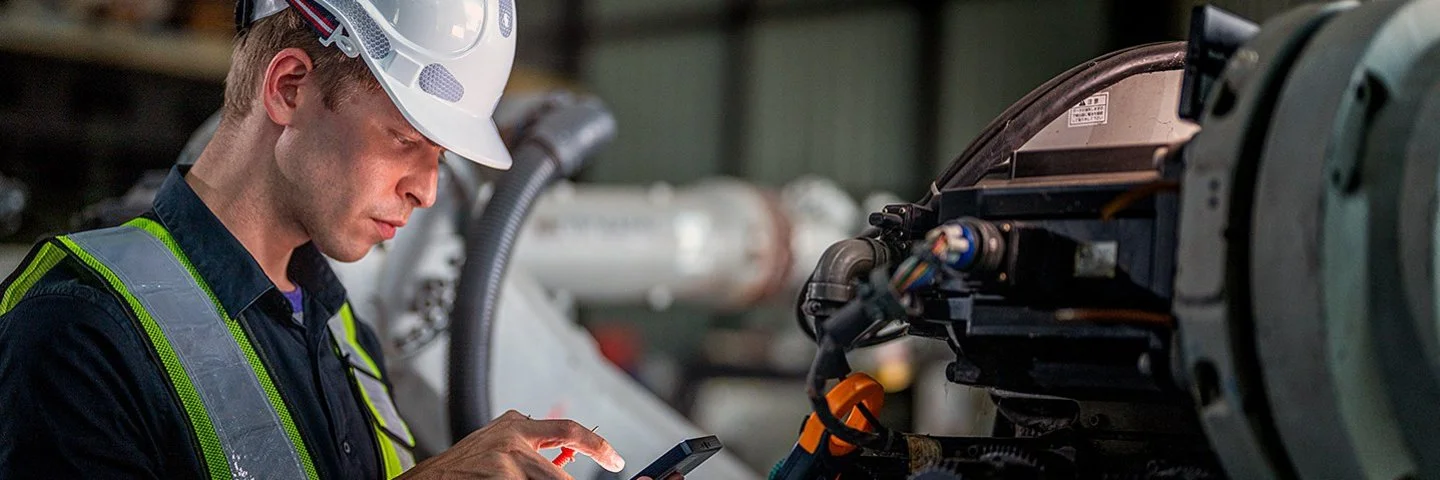 In einer Fabrikhalle steht ein Mann mit einem Schutzhelm vor einer Maschine. Er hält ein elektronisches Wartungs- oder Steuergerät in der linken Hand, blickt auf dessen Display und tippt mit der rechten Hand etwas ein.
