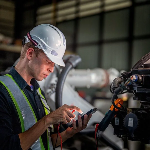 In einer Fabrikhalle steht ein Mann mit einem Schutzhelm vor einer Maschine. Er hält ein elektronisches Wartungs- oder Steuergerät in der linken Hand, blickt auf dessen Display und tippt mit der rechten Hand etwas ein.