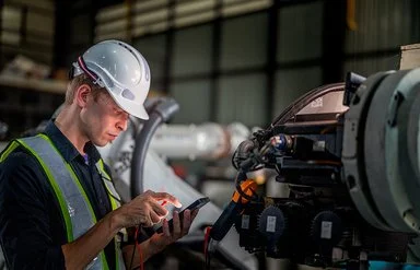 In einer Fabrikhalle steht ein Mann mit einem Schutzhelm vor einer Maschine. Er hält ein elektronisches Wartungs- oder Steuergerät in der linken Hand, blickt auf dessen Display und tippt mit der rechten Hand etwas ein.
