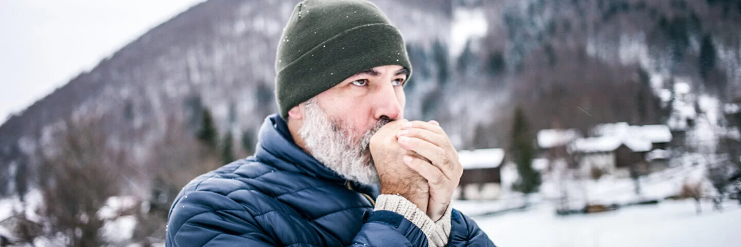 Ein Mann in Winterkleidung hält sich seine kalten Hände vor dem Mund, um sie zu wärmen.