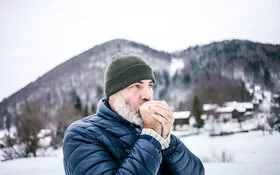 Ein Mann in Winterkleidung hält sich seine kalten Hände vor dem Mund, um sie zu wärmen.