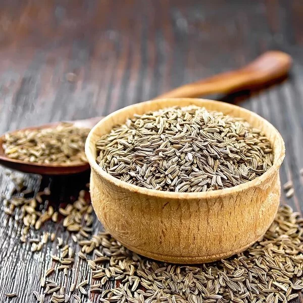 Auf einem Holztisch steht ein Holzschälchen mit Kreuzkümmelsamen. Hinter dem Schälchen liegt ein Holzlöffel, ebenfalls mit Kreuzkümmelsamen. Weitere Samen sind um das Schälchen herum auf dem Tisch verstreut.
