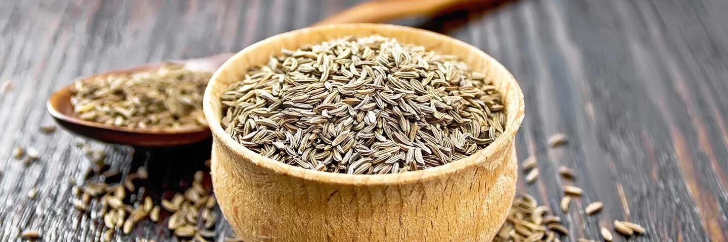 Auf einem Holztisch steht ein Holzschälchen mit Kreuzkümmelsamen. Hinter dem Schälchen liegt ein Holzlöffel, ebenfalls mit Kreuzkümmelsamen. Weitere Samen sind um das Schälchen herum auf dem Tisch verstreut.