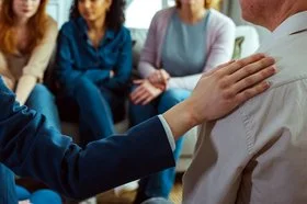 Eine Teilnehmerin einer Selbsthilfegruppe legt einem Mann ihre Hand auf die Schulter.