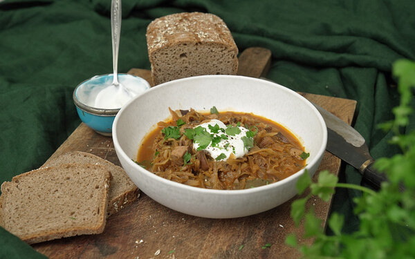 Auf einem Holzbrett steht eine Schüssel vegetarisches Bigos mit Brot und Joghurt.