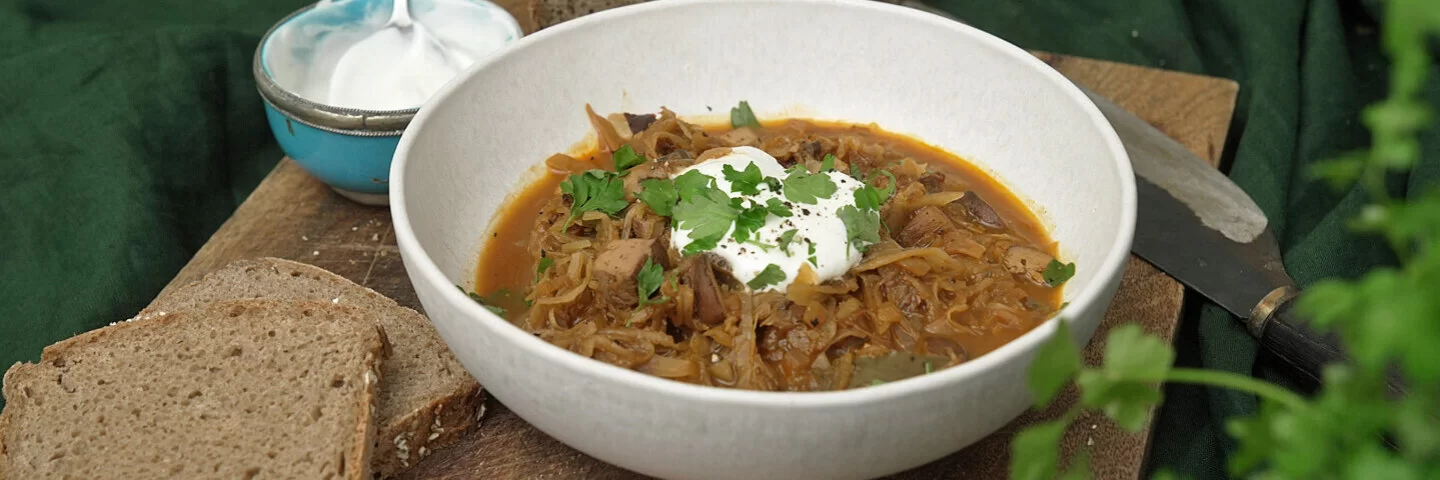 Auf einem Holzbrett steht eine Schüssel vegetarisches Bigos mit Brot und Joghurt.