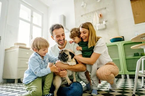 Eine junge vierköpfige Familie hockt auf dem Küchenboden. Auf den Oberschenkeln des Vaters sitzt ein Mops, eines der Kinder streichelt den Hund. Die Mutter und ein jüngeres Kind beobachten die Szene. Alle lachen fröhlich.