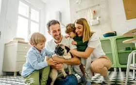 Eine junge vierköpfige Familie hockt auf dem Küchenboden. Auf den Oberschenkeln des Vaters sitzt ein Mops, eines der Kinder streichelt den Hund. Die Mutter und ein jüngeres Kind beobachten die Szene. Alle lachen fröhlich.
