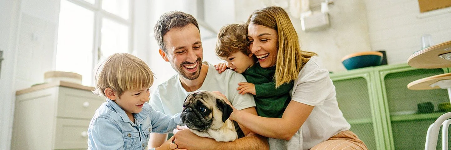Eine junge vierköpfige Familie hockt auf dem Küchenboden. Auf den Oberschenkeln des Vaters sitzt ein Mops, eines der Kinder streichelt den Hund. Die Mutter und ein jüngeres Kind beobachten die Szene. Alle lachen fröhlich.